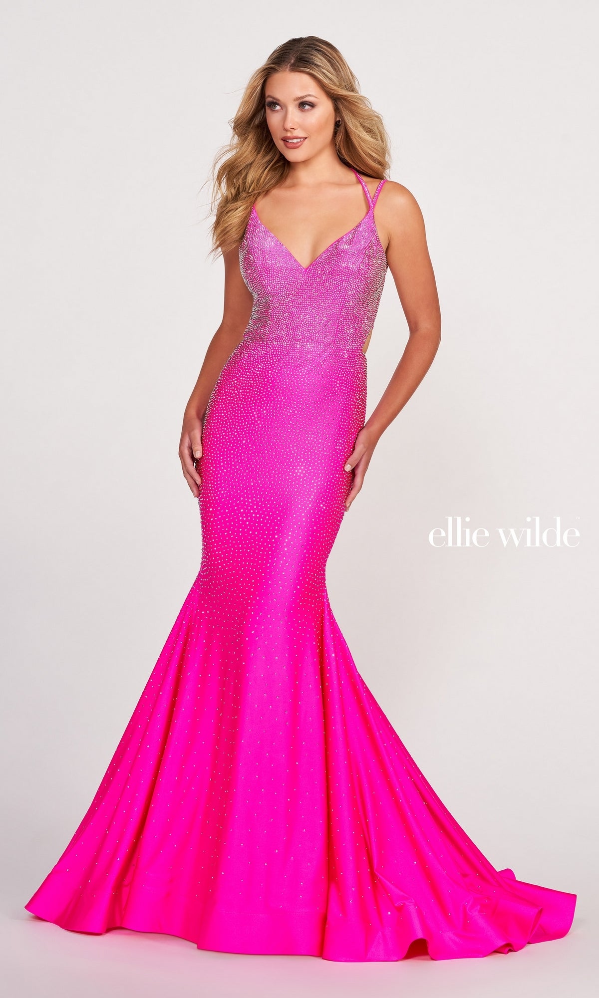 A woman poses against a plain background in the Strappy-Back Long Beaded Ellie Wilde Formal Gown—a bright pink, floor-length dress with thin straps and subtle embellishments. The text